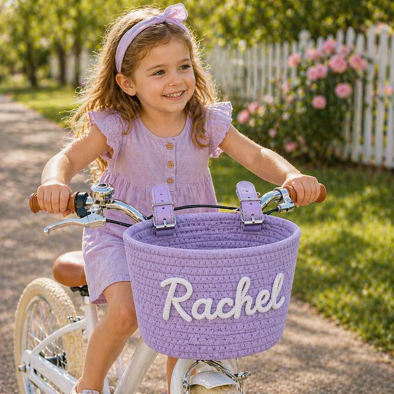 May include: A young girl rides a white bicycle with a lavender basket. The basket is embroidered with the name "Rachel" in white. The girl wears a matching lavender dress and headband. The bike has brown handlebars and a white seat.