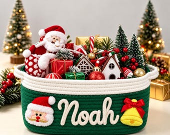 May include: A dark green rope basket with a white rim, featuring a plush Santa face, the name Noah in white script, and a yellow bell with a red bow. The basket is filled with Christmas ornaments, miniature houses, and small pine trees.