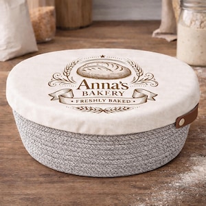 Op de afbeelding: Een ronde, grijs en witte geweven mand met een crèmekleurig deksel. Het deksel heeft een bruin bakkerijlogo met de tekst "Anna's Bakery" en "Freshly Baked". Een bruine leren handgreep is aan de zijkant bevestigd.