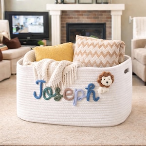 May include: A white rope basket with the name "Joseph" in blue, green, and brown yarn. The basket contains a yellow pillow, a patterned pillow, and a cream-colored blanket. A small lion applique is attached to the basket.