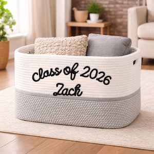 May include: A white and gray woven storage basket with the text "class of 2026 Zach" in black script. The basket contains a beige woven pillow and a gray pillow. The basket is on a light-colored rug.
