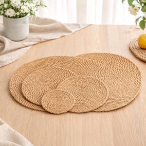 May include: A collection of woven, circular placemats and coasters in different sizes. The natural-coloured placemats and coasters are displayed on a light wooden table. The placemats and coasters are made from a tightly woven material.
