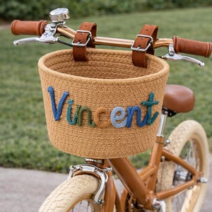May include: A brown bicycle with a woven basket attached to the handlebars. The basket has the name "Vincent" stitched in blue, green, and orange yarn. The bike has brown leather accents and cream-colored tires.