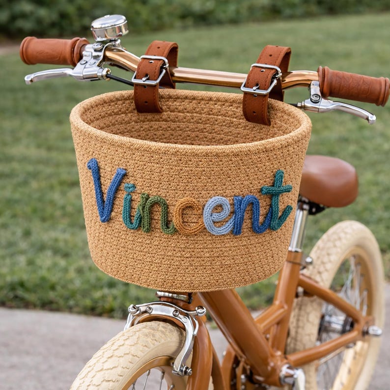 May include: A brown bicycle with a woven basket attached to the handlebars. The basket has the name "Vincent" stitched in blue, green, and orange yarn. The bike has brown leather accents and cream-colored tires.