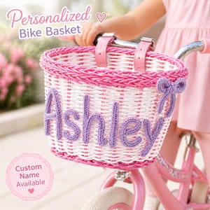 Puede incluir: Una cesta de bicicleta tejida rosa y blanca con el nombre "Ashley" en hilo lavanda. La cesta tiene correas de cuero rosa y un lazo decorativo. El texto en la imagen dice "Cesta de bicicleta personalizada" y "Nombre personalizado disponible."