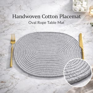 May include: A light grey, handwoven, oval cotton placemat. The placemat is made of braided rope and is displayed with a gold fork and knife. The text "Handwoven Cotton Placemat Oval Rope Table Mat" is at the top.