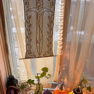 May include: A decorative window treatment featuring a brown macrame panel suspended from a wooden rod, set against sheer, cream-colored curtains. The scene includes a stained glass lamp, plants, and other decorative items.