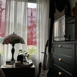Może przedstawiać: Dwie prostokątne szydełkowe zawieszki ścienne z misternymi czerwonymi wzorami mieczy zawieszone w oknie. Lewy element przedstawia pojedynczy duży miecz, podczas gdy prawy element pokazuje miecz rozpadający się na mniejsze odłamki z tekstem.