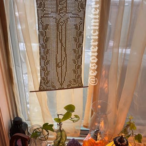 May include: A rectangular beige crochet wall hanging featuring a central sword motif, suspended from a thin wooden rod. The piece displays an intricate openwork pattern and includes the text @esotericinterval vertically along the right side.