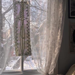 Peut inclure: Une chambre ensoleillée avec une fenêtre encadrée de rideaux en dentelle transparente. Une pièce décorative rectangulaire est suspendue à la fenêtre, avec un motif d'épée. Le lit est recouvert d'une couette à motifs floraux et d'oreillers.