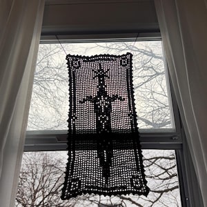 Może przedstawiać: Czarny, szydełkowy element dekoracyjny wisi w oknie. Element jest prostokątny z szczegółowym wzorem miecza pośrodku. Okno jest obramowane białymi zasłonami, a tło przedstawia nagie gałęzie drzew.