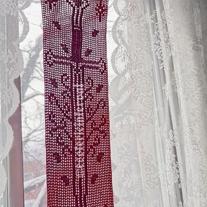 Peut inclure: Une cantonnière de fenêtre en crochet bordeaux avec un motif vertical central et des motifs floraux. La cantonnière est suspendue devant une fenêtre avec des rideaux en dentelle blanche. Le design est symétrique et détaillé.