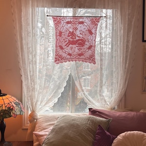 May include: A bedroom scene featuring a window with sheer lace curtains and a red lace valance with a deer design. The bed is covered with floral patterned bedding and various pillows in shades of pink and green.