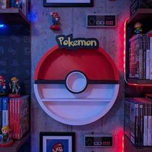 May include: A red, white, and black Pokemon-themed shelf, shaped like a Pokeball, mounted on a gray wall. The wall also features framed pixel art of Mario, Nintendo controllers, and figurines of video game characters. Shelves display video game cartridges and consoles.