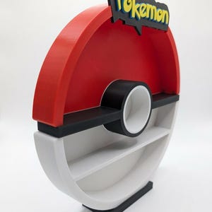 May include: A red, black, and white shelf unit shaped like a Pok&eacute; Ball. The top features the word "Pokemon" in yellow and blue. The shelf has two interior shelves and a black base.