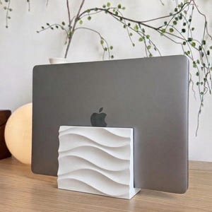 May include: A white rectangular laptop stand featuring a textured, wavy, three-dimensional surface pattern on the front. The stand is holding a dark gray laptop in an upright, vertical position, displaying the metallic Apple logo on the back.
