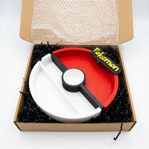 May include: A round, divided tray in a cardboard box, designed to resemble a Pok&eacute; Ball. The tray is red and white with a black stripe and a black and yellow "Pokemon" logo. The box is filled with black shredded paper.