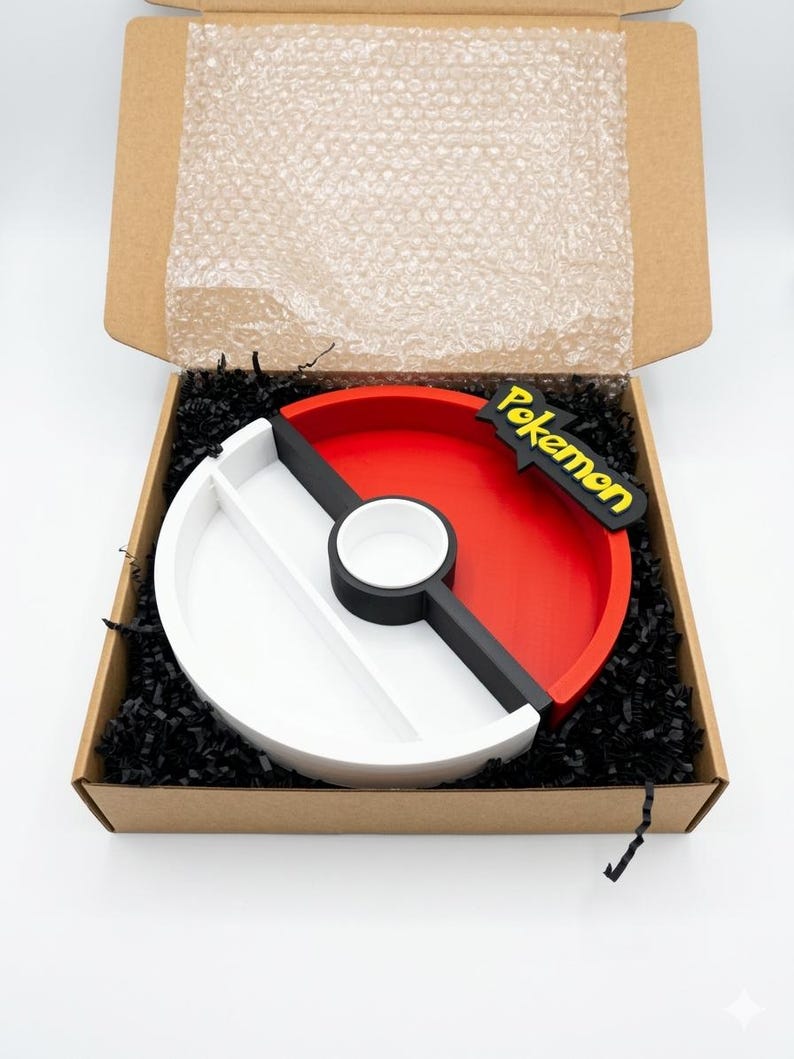 May include: A round, divided tray in a cardboard box, designed to resemble a Pok&eacute; Ball. The tray is red and white with a black stripe and a black and yellow "Pokemon" logo. The box is filled with black shredded paper.