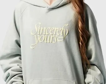 May include: A pale green hoodie with a kangaroo pocket and a hood. The words "Sincerely yours" are embroidered in a cursive font across the chest. The sleeves are long and the cuffs are ribbed.
