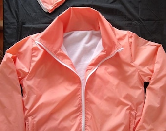 May include: A peach-colored windbreaker jacket with a white front zipper closure, a high collar, and long sleeves with elastic cuffs. The jacket features a white interior lining and is accompanied by a matching detachable hood.