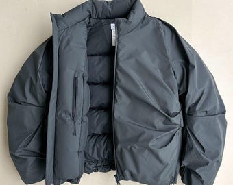 May include: A dark grey puffer jacket with a high collar and a full-length zip. The jacket has a streamlined design with a subtle texture and a small zipped pocket on the chest. The jacket is open, revealing the quilted interior.