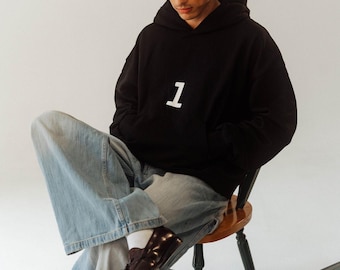 May include: Black hoodie with a white "1", worn with light blue jeans. The person is seated on a wooden chair, wearing brown boots. A vintage suitcase is next to the chair.