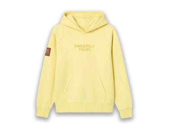 May include: A pale yellow hoodie featuring a hood and a front pocket. The words "SINCERELY YOURS" are printed on the front. A small patch is on the left sleeve. The hoodie is made of a soft material.