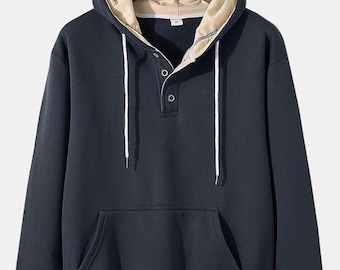 May include: A dark navy blue long-sleeve hoodie featuring a beige inner hood lining, a front placket with two silver-toned metal snap buttons, and white drawstring cords. The garment includes a large front kangaroo pocket and ribbed cuffs.