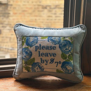 May include: A light blue decorative pillow with a cross-stitched design. The cross-stitch features blue roses and the text "please leave by 7#". The pillow has a light blue border and is resting on a wooden surface.