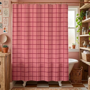 May include: A pink shower curtain with a grid pattern in a darker shade of pink. The curtain hangs from a copper-colored rod in a bathroom setting. The bathroom has a white bathtub, wooden shelves, and a window.