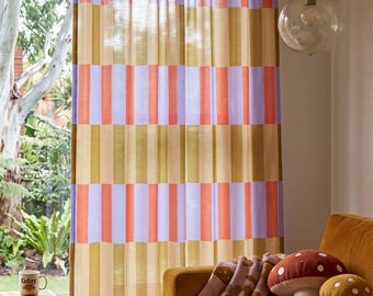 May include: A curtain featuring a vertical striped pattern in shades of lavender, orange, and mustard yellow. The fabric is semi-sheer, allowing light to pass through. The design consists of repeating rectangular blocks of colour arranged in columns.