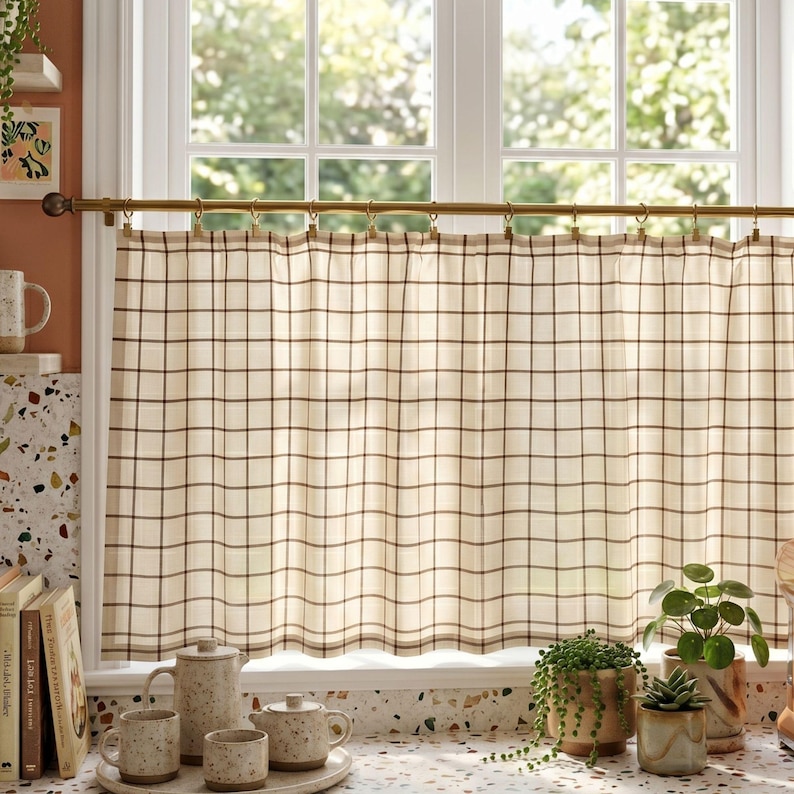 May include: A cafe curtain featuring a cream-colored fabric with a dark brown grid pattern. The curtain is suspended from a brass-toned metal rod using small metal clips. The fabric is semi-sheer, allowing light to pass through the window pane.