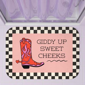 May include: A rectangular bath mat with a black and white checkered border and a pink center. The design features a red cowboy boot with pink flame details and a small heart, accompanied by the black text GIDDY UP SWEET CHEEKS.