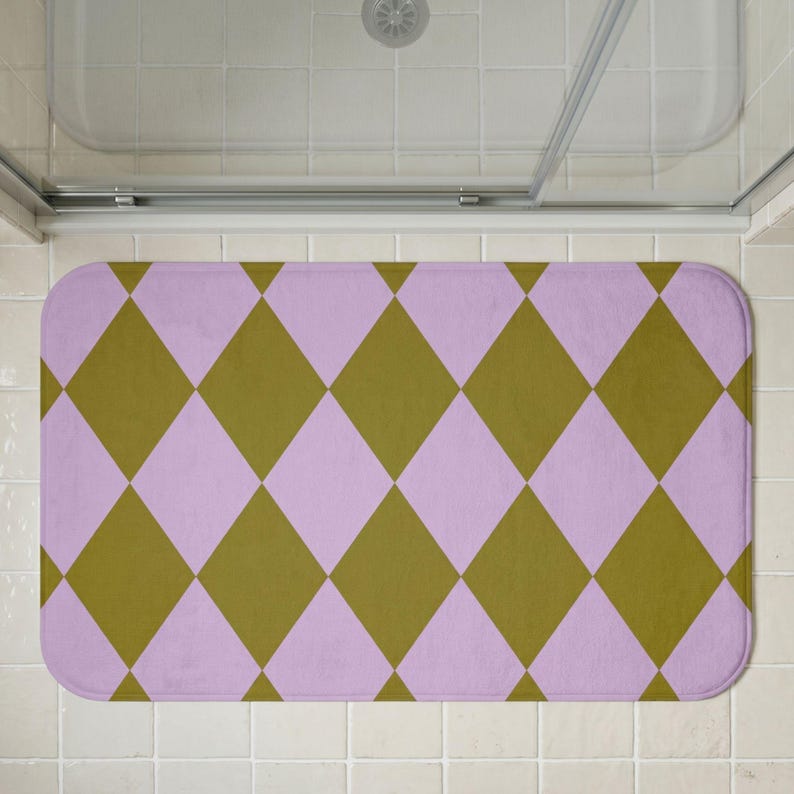 May include: A rectangular bath mat featuring a repeating harlequin diamond pattern in alternating shades of muted olive green and soft pastel lavender. The mat has rounded corners and a flat, uniform texture across the entire surface area.
