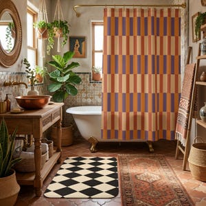 May include: A shower curtain featuring a repeating vertical pattern of alternating orange, cream, and purple rectangular blocks. The curtain hangs from a gold-toned rod and covers a white clawfoot bathtub in a bathroom with tiled walls and floors.