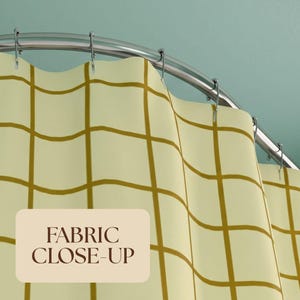 Pu&ograve; includere: Una tenda da doccia con un motivo a griglia a linee giallo senape su sfondo color crema pallido, appesa a un'asta curva in metallo argentato con anelli. Un'etichetta beige riporta il testo FABRIC CLOSE-UP.