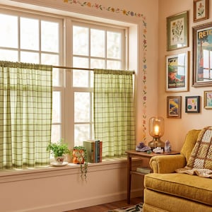 May include: A pair of light green cafe curtains with a dark green grid pattern hanging on a thin metal rod over a window. The curtains are semi-sheer and feature a simple rod pocket header, providing light filtration while maintaining privacy.