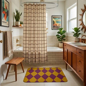May include: A rectangular bath mat featuring a repeating diamond pattern in alternating shades of mustard yellow and deep purple. The mat has rounded corners and lies flat on a tiled floor in front of a wooden vanity and a bathtub with a grid curtain.