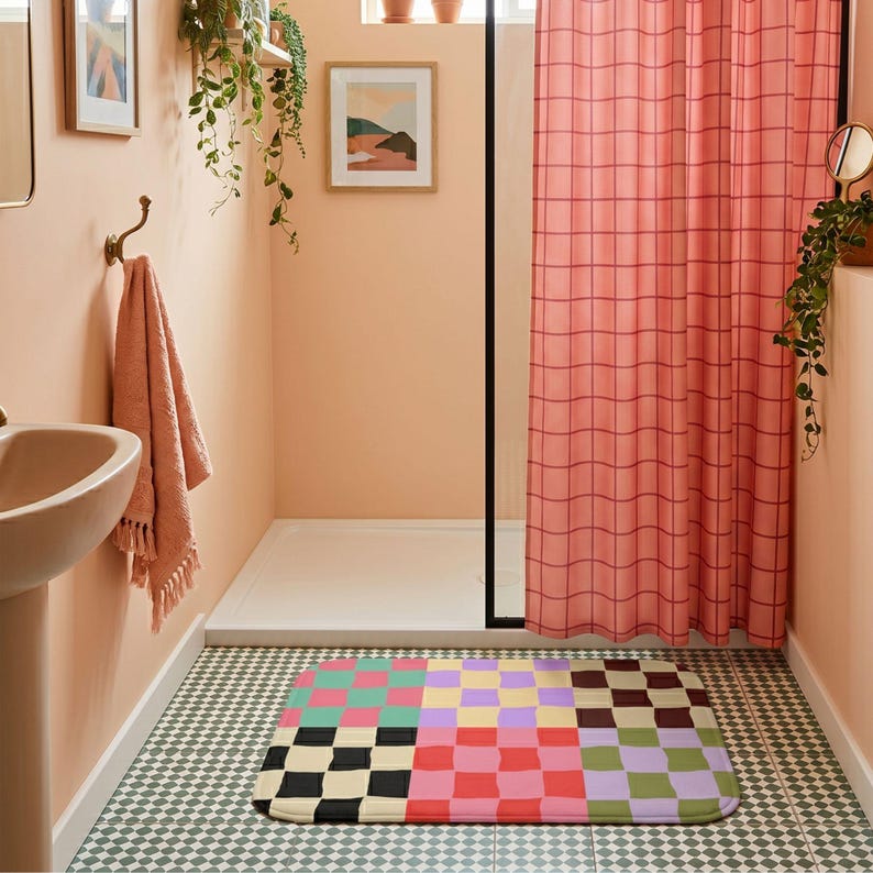 May include: A rectangular bathroom floor mat featuring a multi-colored checkerboard pattern with squares in shades of pink, teal, yellow, purple, black, cream, red, and green, positioned on a tiled floor in front of a pink grid shower curtain.