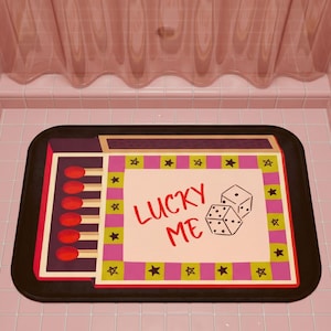 May include: A rectangular bath mat with a black border featuring a graphic of a matchbox on the left side with six red-tipped matches. The center has a cream-colored rectangle with the red text LUCKY ME and two dice, framed by a pink and green border with black stars.