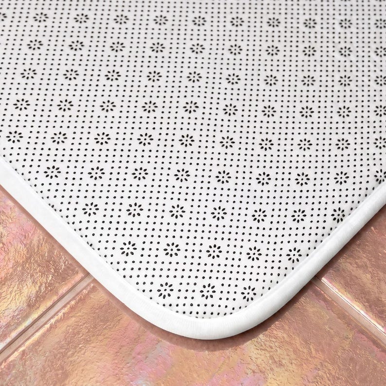May include: A white rectangular floor mat featuring a repeating pattern of small black flower-shaped dots across the surface. The mat has a smooth white fabric border and a textured underside designed for grip on flat surfaces.