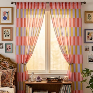 Peut inclure: Une paire de rideaux de fenêtre transparents présentant un motif à blocs verticaux dans des tons alternés de corail, rose clair, jaune et lavande. Les rideaux sont suspendus à une tringle en métal foncé et sont tirés pour laisser passer la lumière.