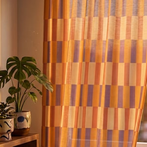 Puede incluir: Cortina transparente con un patrón geométrico de rayas verticales en tonos naranja, amarillo y morado. La tela ligera permite el paso de la luz. A la izquierda, se observa una planta en maceta. El diseño es moderno.