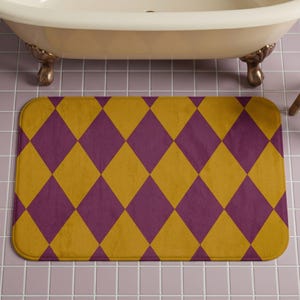 May include: A rectangular bath mat with a repeating harlequin pattern of alternating purple and gold diamonds. The mat is placed on a pink tiled floor beneath a white bathtub with gold claw feet.