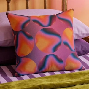 May include: A square throw cushion featuring a repeating pattern of large circles. The design displays a vibrant gradient effect, transitioning through shades of orange, pink, purple, and blue. The fabric has a smooth texture and a matte finish.