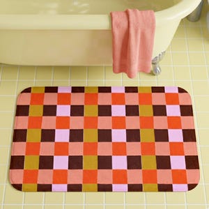 May include: A rectangular bath mat featuring a repeating checkered pattern in shades of dark brown, mustard yellow, bright orange, and light pink. The mat has rounded corners and a flat, uniform texture, positioned on a light yellow tiled floor.