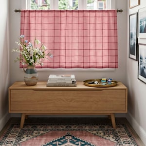 May include: A pink and red plaid window curtain hangs on a rod. Below, a light wood console table holds a ceramic vase with white and blue flowers, a stack of three books, and a gold tray containing keys and small decorative pins.