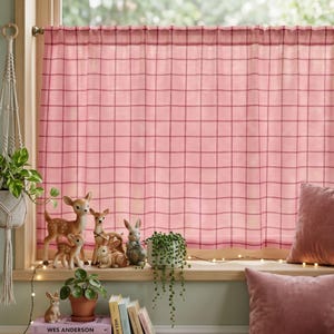 May include: A pink and red plaid cafe curtain hangs on a rod over a window. On the windowsill sit several deer and rabbit figurines, a small potted plant, and a book titled Wes Anderson. A string of fairy lights is draped across the sill.