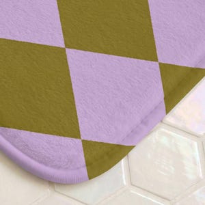 May include: A soft, rectangular bath mat featuring a repeating argyle pattern in alternating shades of light lavender and olive green. The fabric has a plush, textured surface with rounded corners and a finished edge in matching lavender.