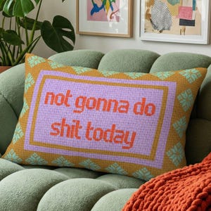 May include: Rectangular decorative pillow with the text "not gonna do shit today" in orange letters on a lavender background. The pillow has an orange border with teal floral accents, resting on a green sofa.